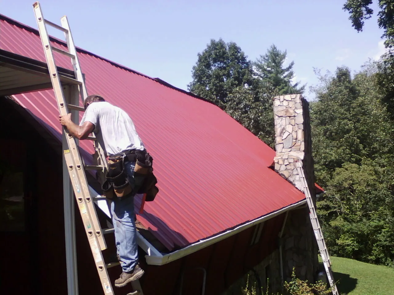 Roofer on a ladder beside a red metal roof and stone chimney — Soffit & Fascia Repair by Roofing Brothers in Amberson